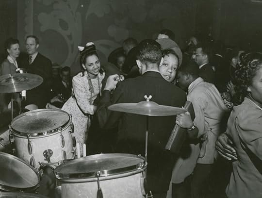 A vintage black-and-white photograph of a lively dance scene featuring couples dancing closely, with a drummer in the foreground and people enjoying the music in a social setting.