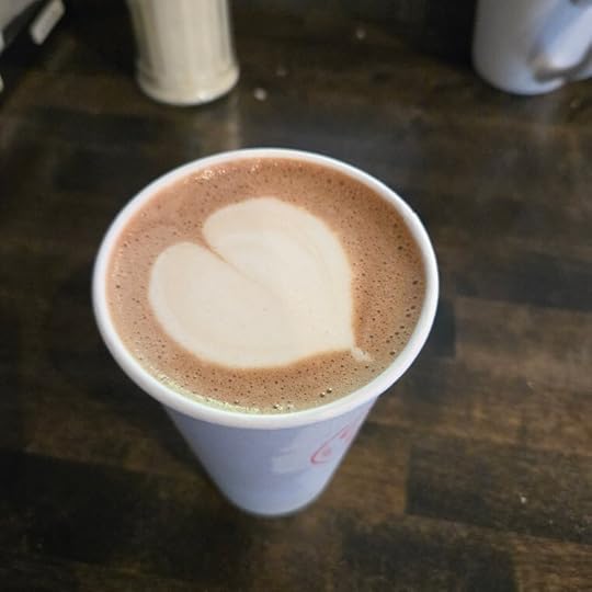 A paper cup of coffee with the foam on top in the shape of a heart sits on a wooden counter. Canisters of sugar and milk can be seen in the background.