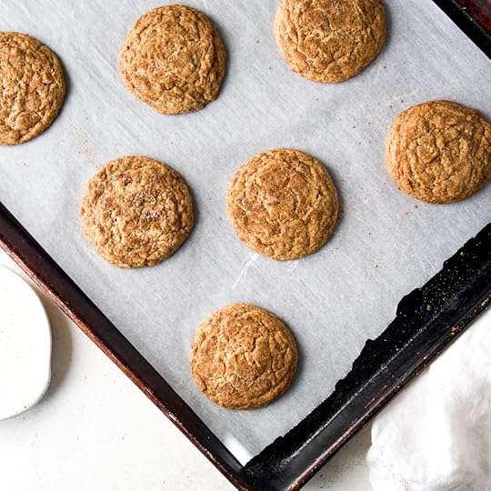 Seven freshly baked, golden brown chai snickerdoodle cookies resting on on parchment paper on a dark metal baking sheet. The cookies show a cracked, soft texture with a spiced sugar coating.