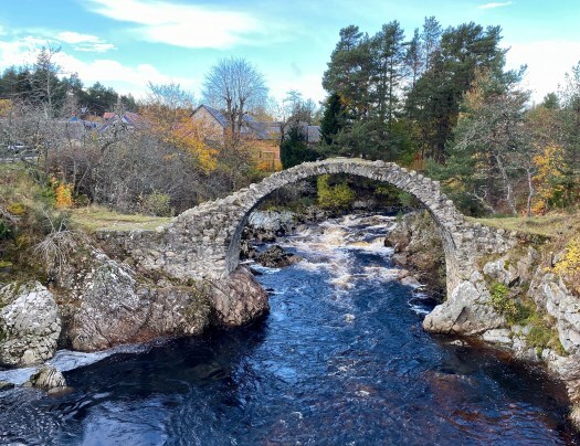 The Coffin Bridge of Carrbridge over a rough dark-blue river, autumnal trees behind