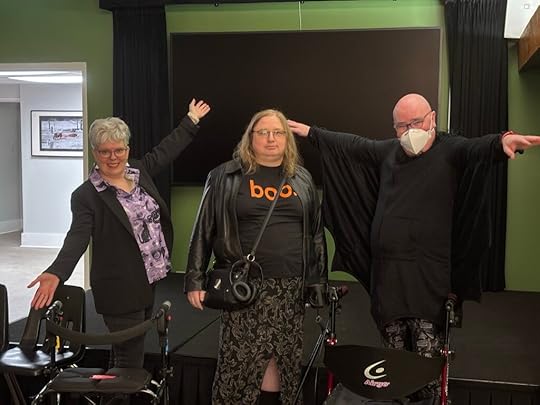 Talia is in the middle, all in black except for the lowercase “boo” in orange on her T-shirt. Derek is on the right in this photo, wearing a cloak that looks like bat wings with their arms stretched. I’m on the left, in a black blazer and purple shirt with halloween creatures on it. Both Derek and I use rollators.