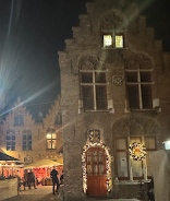A building against a night's sky. The red door has a fairy lit garland around it. The window has a large illuminated star dangling. To the side of the building, in the distance, is a market square. The fairy lights are strung across outdoor umbrellas outside the restaurants.