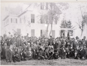 A historical black and white photograph of a large group of men gathered outdoors, some wearing hats and holding walking sticks, with a building in the background.