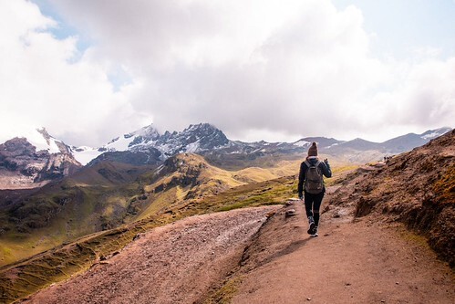 Hiker walking along a mountain trail toward snow-capped peaks under a cloudy sky in Peru.