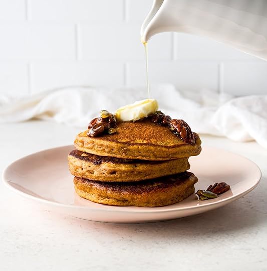 Stack of three fluffy homemade pumpkin pancakes on a pink plate, topped with a pat of butter, candied pecans, pumpkin seeds and real maple syrup being poured from a white pitcher.
