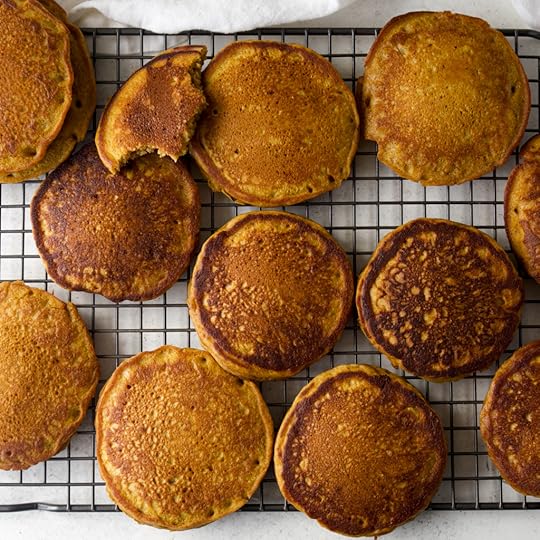 Overhead view of a large batch of finished, golden-bowl pumpkin pancakes cooing on a wire rack, with one pancake showing the fluffy interior texture.