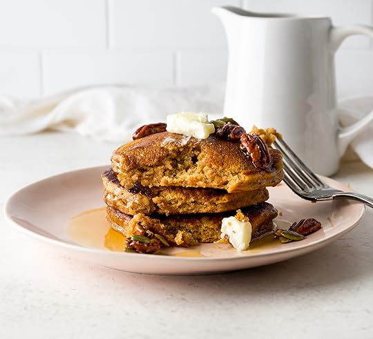 Stack of three pumpkin pancakes on a pink plate with a bite taken out of the one on top showing a moist, airy texture. Topped with butter, maple syrup, pecans and pumpkin seeds.