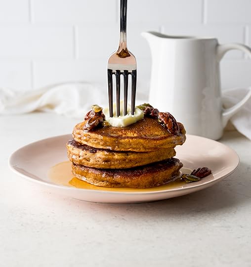Stack of three fluffy homemade pumpkin pancakes on a pink plate drizzled with maple syrup and topped with pecans and pumpkin seeds. A silver fork is stuck into the top pancake.