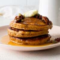 Stack of fluffy homemade pumpkin pancakes topped with butter, maple syrup, pecans and pumpkin seeds on a pink plate.