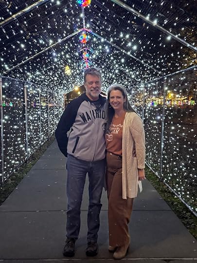 A man and a woman standing together under a canopy of twinkling holiday lights, smiling as they pose for a photo.