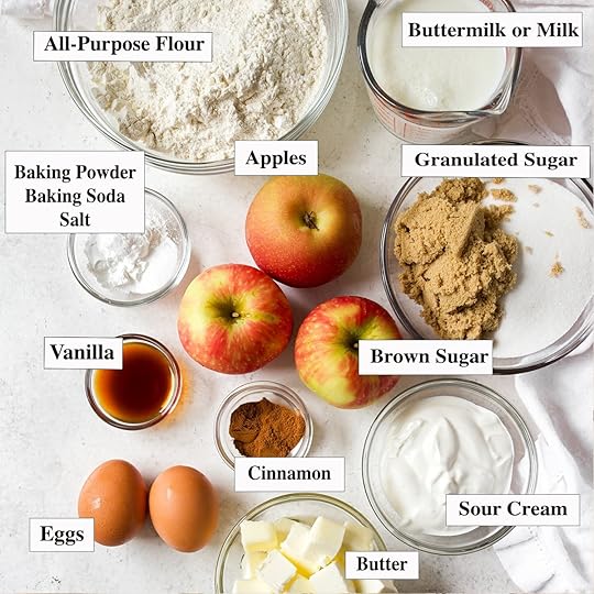 Overhead view of all of the ingredients for mega moist apple muffins laid out, including apples, flour, brown sugar, sugar, butter, sour cream, buttermilk, eggs, vanilla and spices.