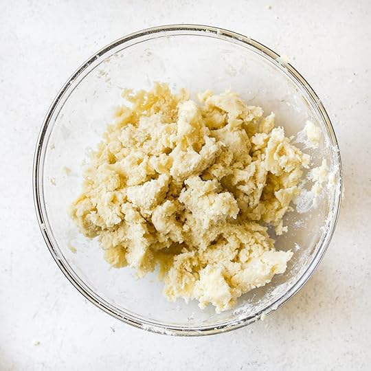 Close-up overhead view of blonde streusel topping with a crumbly, shaggy texture in a clear glass bowl, ready to top cinnamon apple muffins.