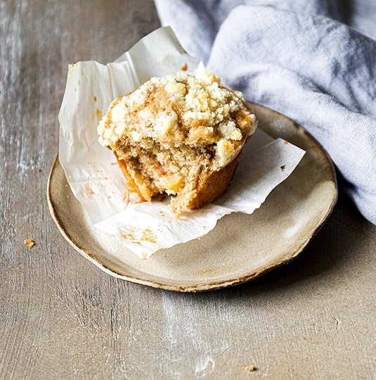 Single mega moist apple cinnamon muffin with a streusel crumble topping, a bite taken out, served on a rustic tan plate with a crumpled blue linen napkin in the background.