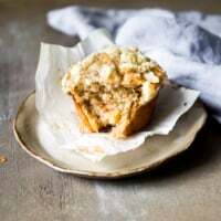 Mega moist apple cinnamon muffin with streusel topping and a bite taken out, on a plate with parchment liner, next to a blue linen napkin.