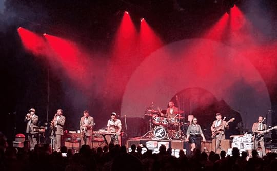 A live performance by Lake Street Dive, featuring the band members playing various instruments on stage with dramatic red lighting and a crowd visible in the foreground.