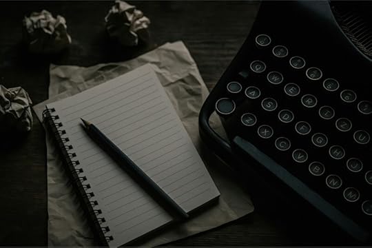 A vintage typewriter next to a spiral notebook with a pencil, surrounded by crumpled papers on a wooden table.
