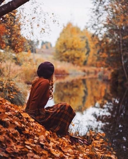 This may contain: a woman sitting on the ground next to a tree and water in an autumn forest