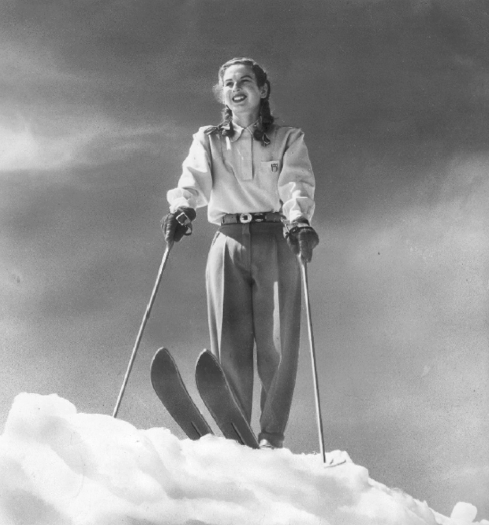 A female figure skater with short hair smiles and skates outdoors, arms outstretched. Another blurred skater is in the background.