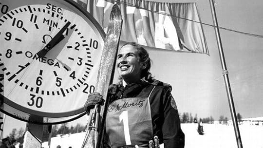 A female figure skater with short hair smiles and skates outdoors, arms outstretched. Another blurred skater is in the background.