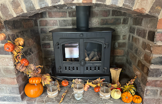 A stone fireplace. Around the hearth, there's a garland of orange leaves and pinecones. Sitting side are a selection of pumpkins in orange, green, and pale white. The pale white one has a large brown parch where it's started to rot.