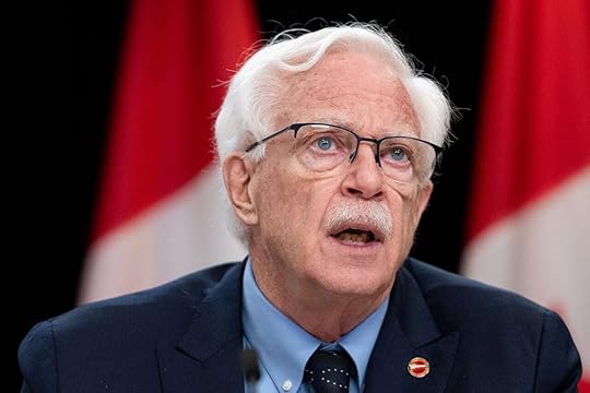 A man in a suit, wearing glasses, speaks to someone off screen. Canadian flags are seen in the background.