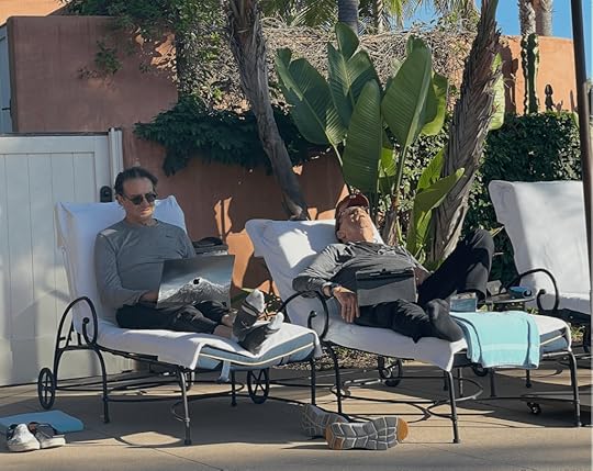 Two men lounging by a poolside on sunbeds; one is working on a laptop while the other is relaxing with his eyes closed.