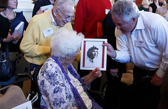 At a crowded event, two smiling men show a seated woman a photograph of herself as a young woman. 