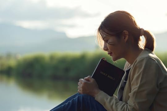 Young woman praying outdoors while holding a Bible beside a quiet lake at sunset.