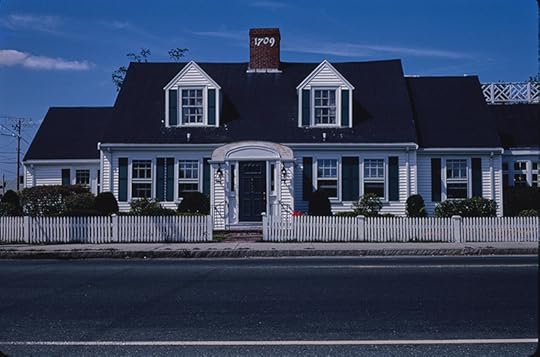 Landscape style photograph of a traditional two-story home with two dormer windows. It is a wide, white home with a black roof and shutters and a white picket fence in front. 
