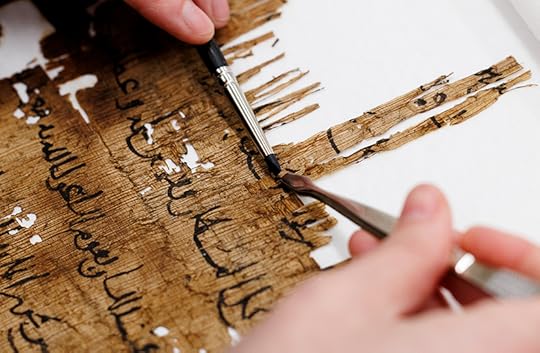 A close up of a ragged sheet of browned, brittle papyrus with writing in black ink. A conservators hands are holding two thin metal instruments at one point on the papyrus.