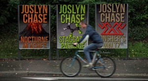 Man riding a bicycle past a wall of giant posters showing the covers of the first three books of the Riley Forte Suspense Thrillers, by Joslyn Chase