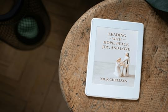 A white e-reader displaying the cover of Leading with Hope, Peace, Joy, and Love by Nick Chellsen, resting on a rustic wooden table next to a soft light source. The cover features a minimalist nativity scene with Mary, Joseph, and baby Jesus.