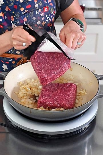 Someone in a blue apron is adding two packages of raw ground beef to a large gray skillet filled with sautéed diced onions.