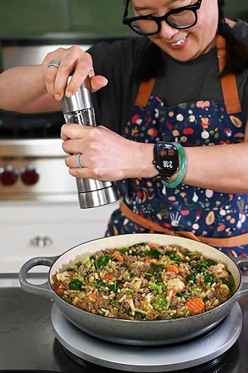 A smiling Asian woman in glasses and braids is adding freshly cracked black pepper from a pepper mill into a large gray skillet with cooked ground beef, shredded potatoes, and mixed vegetables.