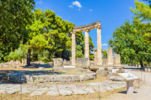Photograph of ruins of Ancient Olympia, Greece shows three columns remain standing at the edge of a circular patio of stones with green trees in the background