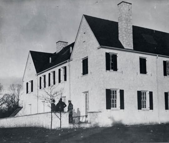 Black and white photo of a large two-story stone building with multiple windows and a sloped roof, featuring several people standing outside near a low wall.