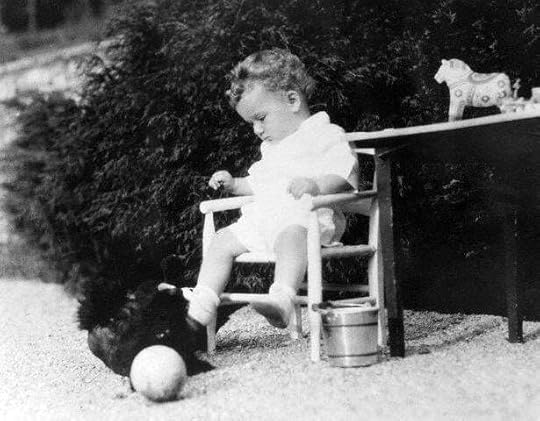 A black and white photo of a young child sitting in a small chair, playing with a toy dog and a ball, next to a small table with a toy horse. The child is dressed in a white outfit and appears focused on their play.