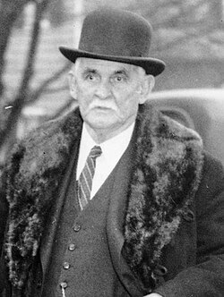 Black and white photograph of an elderly man wearing a bowler hat and a long coat with a fur collar, standing outdoors with a serious expression.