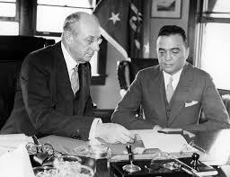 Two men engaged in a serious discussion at a desk, reviewing documents, with a window and flags visible in the background.