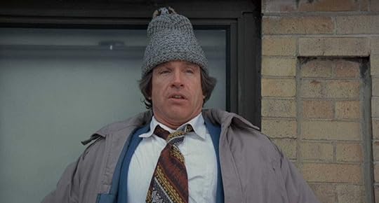 Warren Beatty stands on the ledge of a building with a red knitted beanie on his head