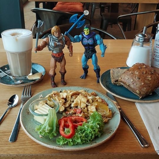 Masters of the Universe Classics Battle Armour He-Man and Skeletor pose with scrambled eggs with vegetables, a slice of bread and a roll and a latte macchiato.