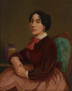 Portrait of Christiana Carteaux Bannister by Edward Mitchell Bannister shows a young woman sitting profile in a green button tufted chair, her right elbow lies on a side table, her hands are clasped, her face is turned slightly toward the viewer. She has center-parted dark hair in an updo, and wears a brown Spencer jacket over a white collared shirt, with a red bow at her throat. Her skirt is full and a slightly lighter shade than her bow.