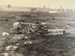 Black and white image of a crashed helicopter in a grassy area with pieces of wreckage scattered around, and several people inspecting the site.