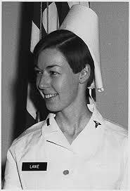 A young female nurse in uniform, smiling, with a name tag that reads 'LANE'. She has short hair and is positioned next to a flag.