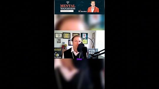 Podcast host seated in a home studio speaking into a microphone with framed awards and a sign in the background.