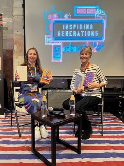 Two women sitting on chairs in front of a colorful backdrop with the text 'Inspiring Generations'. One woman holds two books, and the other holds a book and a notebook. A small table with two water bottles is in front of them.