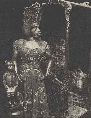 A vintage photograph of a mannequin dressed in a floral gown standing beside a small doll, with a mirror in the background.