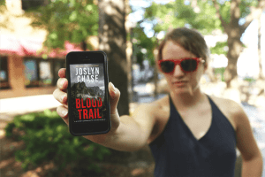 Woman wearing red sunglasses holds out her phone with Cincher's Waltz by Joslyn Chase on the screen