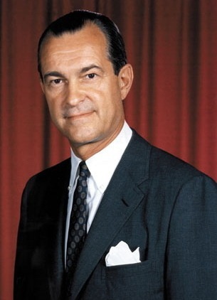 A well-dressed man in a dark suit and tie, smiling against a red curtain background.