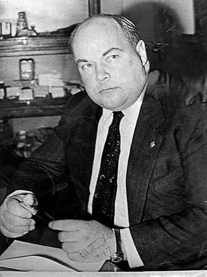 A black and white portrait of a serious man sitting at a desk, holding a pen and looking directly at the camera.